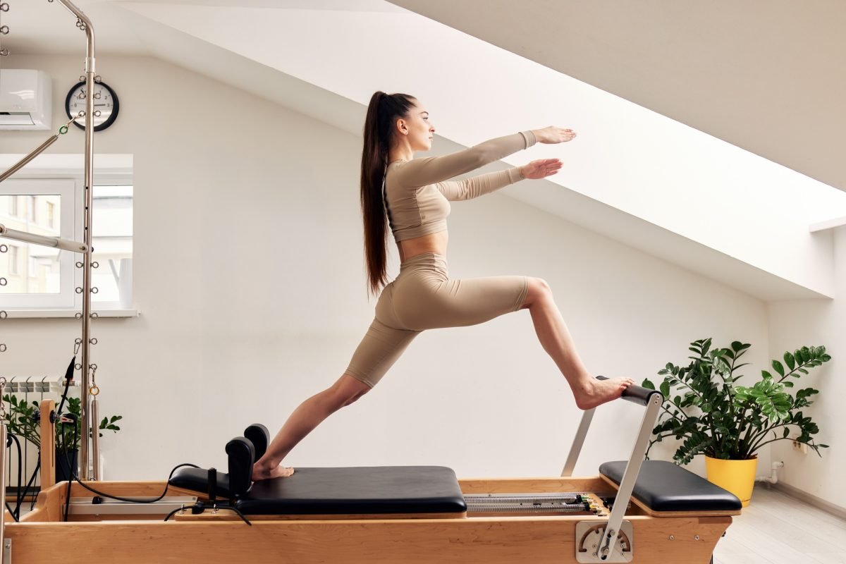 A young girl is doing Pilates on a reformer bed in a bright studio. A slender brunette in a beige